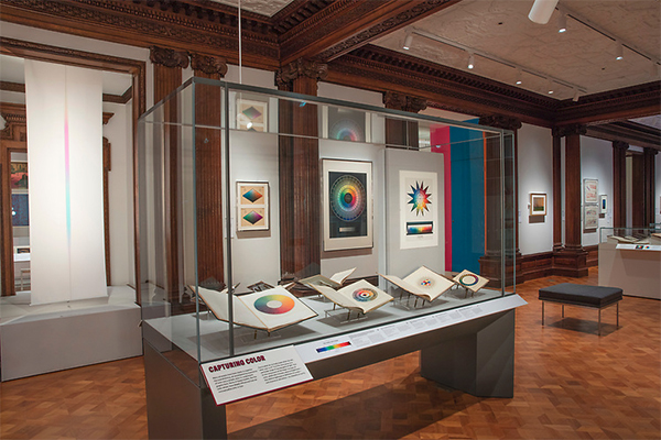 Color books on display in a case with color charts and graphs on the wall in the background at Cooper Hewitt Museum